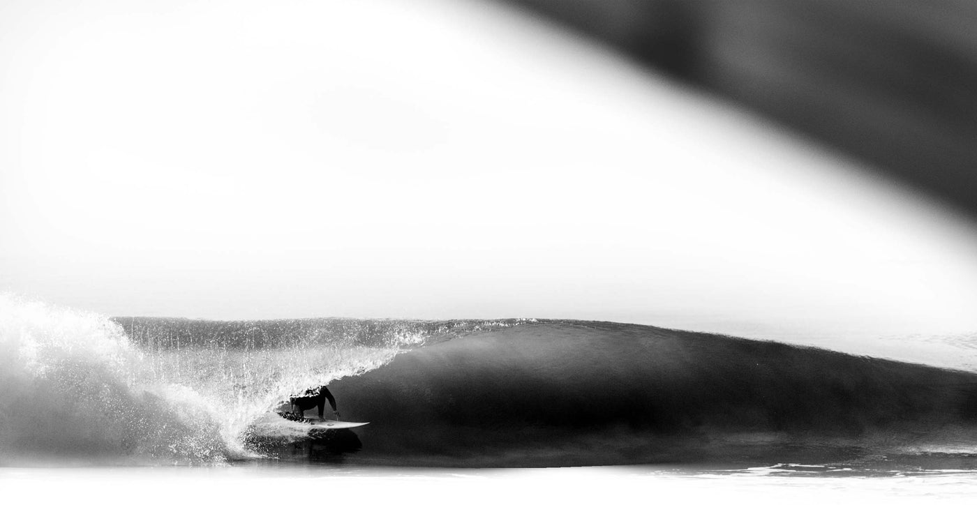 A surfer riding inside the tube of a wave in Indonesia, the surfer is going left on his back side, on a 5'6" FLKLR Surfboard. The image is in Black and white. 